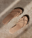 Two tan Eve Sandals with thin, minimalist crisscrossing nappa leather straps rest on a textured beige surface in natural light. Joan Kynyc is embossed on the footbeds, and soft shadows are cast around them.