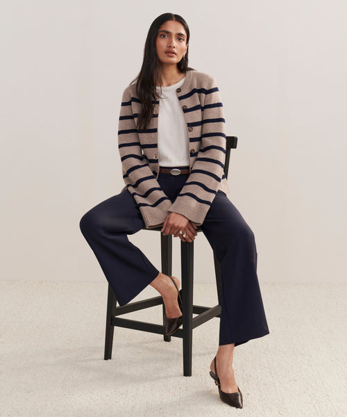 A woman with long dark hair sits on a tall black chair, wearing the Cooper Cardigan in taupe/navy stripe over a white top, navy wide-leg pants, and brown shoes. She faces forward with a neutral expression.