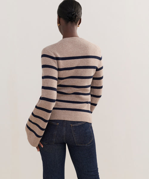A person with hair in a low bun faces away, wearing the Cooper Cardigan in Taupe/Navy Stripe, featuring wide horizontal stripes, a structured silhouette, and slightly flared sleeves, paired with dark blue jeans.