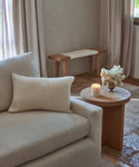 A cozy living room with a light beige sofa, the Nell Boucle Lumbar Pillow in ivory, a round wooden side table with a lit candle and white flowers, stone tile flooring, and neutral curtains.