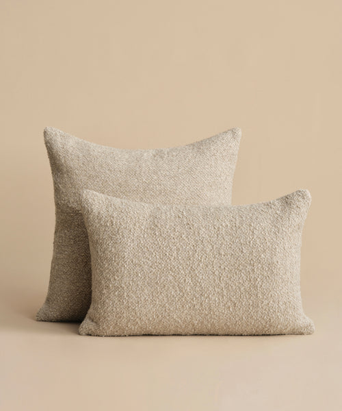 Two Nell Boucle Pillows with feather down inserts are arranged on a light beige background, with one natural, rectangular, textured pillow slightly overlapping the other.