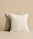 The Nell Boucle Pillow is featured in front—a white, textured square pillow—paired with a light beige natural pillow in back, both set against a beige background.