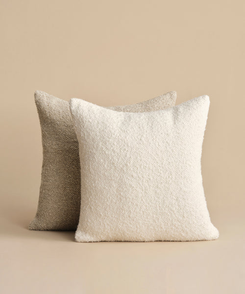 The Nell Boucle Pillow is featured in front—a white, textured square pillow—paired with a light beige natural pillow in back, both set against a beige background.