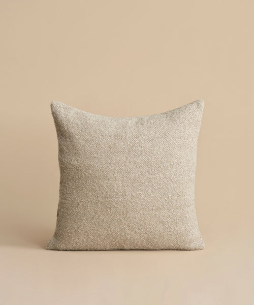 The Nell Boucle Pillow, a beige square pillow with a textured finish, is displayed against a matching background. Its minimalist design is natural and simple, with no visible patterns or embellishments.