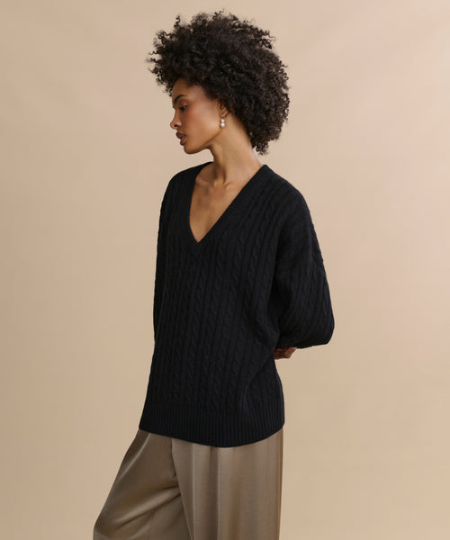 A woman with curly hair wears the Nell V-Neck Sweater—loose, black, cable knit—paired with wide-leg beige pants. She stands against a beige background, arms behind her back, looking to the side.