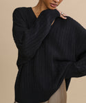 A person poses against a beige background wearing the Nell V-Neck Sweater, a loose-fit black cable knit with long sleeves and ribbed cuffs. Their face is partially out of frame as they hold the edge of the cozy sweater.