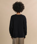 A person with curly hair is seen from behind, wearing the black Nell V-Neck Sweater and beige pants, standing against a plain beige background.