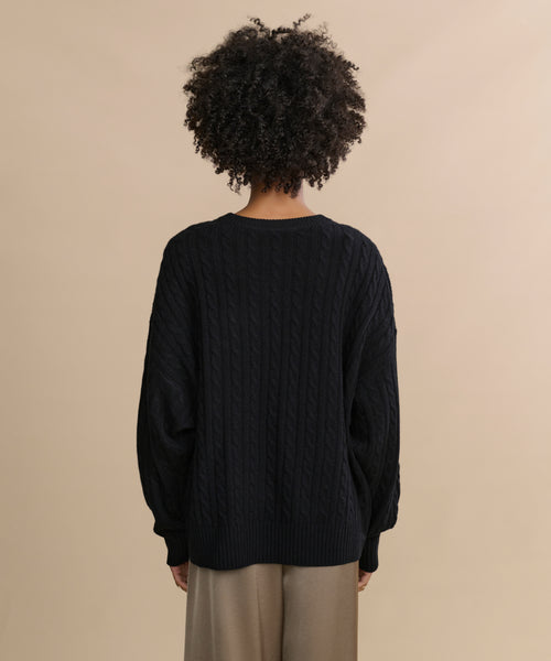 A person with curly hair is seen from behind, wearing the black Nell V-Neck Sweater and beige pants, standing against a plain beige background.