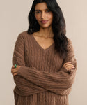 A woman with long, wavy dark hair stands against a neutral background, arms crossed and smiling gently at the camera, wearing the relaxed fit Nell V-Neck Sweater.