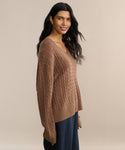 A woman with long dark hair wears the Nell V-Neck Sweater, a relaxed fit brown cable-knit, paired with dark pants. She stands sideways against a beige background, smiling slightly.