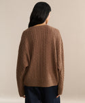A person with long dark hair, seen from behind, wears the Nell V-Neck Sweater in brown and dark pants while standing against a neutral beige background.