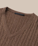 Close-up of the Nell V-Neck Sweater, a brown cable knit with ribbed edges and relaxed fit, displayed on a beige background. A black label is visible inside the V-neck collar.
