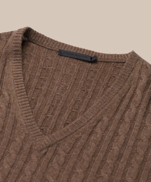 Close-up of the Nell V-Neck Sweater, a brown cable knit with ribbed edges and relaxed fit, displayed on a beige background. A black label is visible inside the V-neck collar.