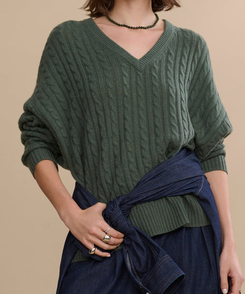 Wearing the Nell V-Neck Sweater in green with a relaxed fit, paired with a blue denim jacket tied at the waist, rings, and a beaded necklace, someone stands against a beige background.