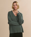 A woman with a blonde bob hairstyle models the Nell V-Neck Sweater in forest green, styled with a simple black necklace, standing against a plain beige background with one hand resting on her shoulder.