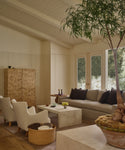 A cozy, neutral living room with a large plant, beige sofa with dark cushions, two armchairs, marble coffee table, and a handwoven Nesting Basket, all warmly lit by natural light from tall windows.