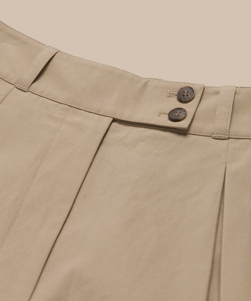 Close-up of the Newport Short in beige Italian cotton poplin, featuring pleats, belt loops, and two brown waistband buttons, set against a matching beige background.