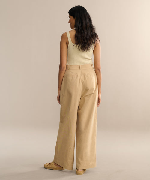 A woman with long, wavy dark hair stands facing away in a cream ribbed tank top and the Newport Trouser in khaki. She pairs this look with tan shoes against a plain, softly lit background.