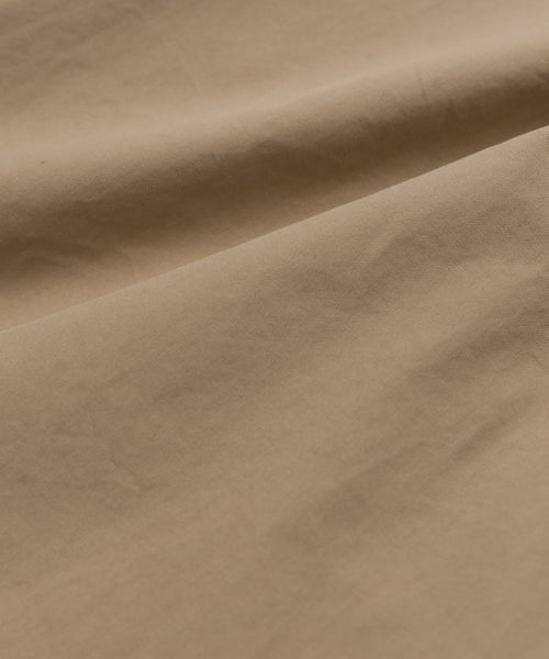 A close-up of smooth, light brown khaki fabric with gentle folds and soft shadows highlights the texture and subtle creases of the Italian cotton poplin featured in our Newport Trouser.
