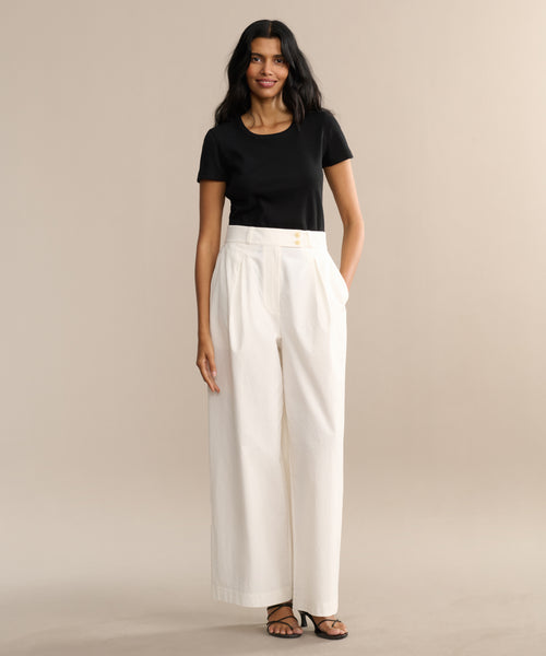 A woman with long dark hair stands against a beige background, wearing a black short-sleeve top, the Newport Trouser, and black sandals. She has a relaxed and confident expression.