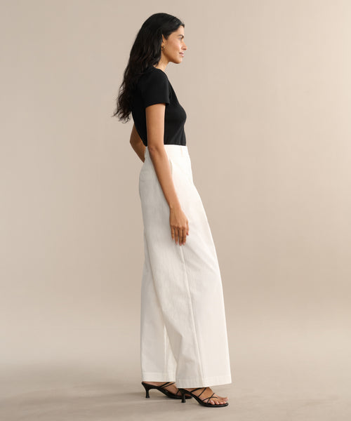 A woman in profile against a beige background wears a black short-sleeve top and high-waisted Newport Trouser, completing her look with black heeled sandals, long dark hair, and a neutral expression.