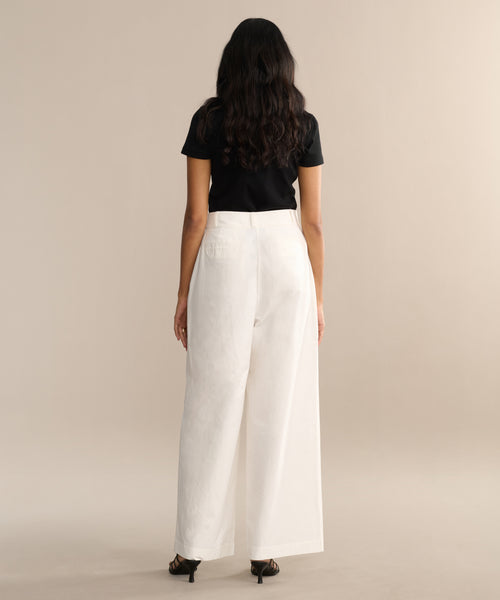 A woman with long dark hair stands facing away, wearing a black short-sleeve top, white Newport Trouser, and black heeled sandals against a beige background.