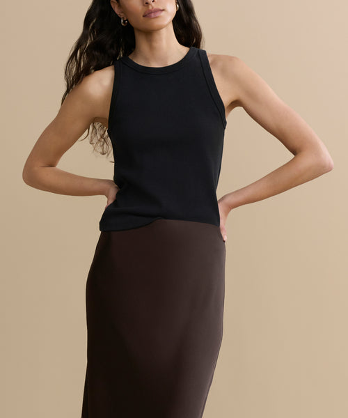 A woman with long, wavy hair wears a sleeveless black top and the high-waisted Nina Slip Skirt in dark brown. She poses against a plain beige background with her hands on her hips.