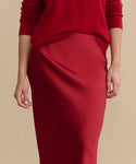A person wearing the Nina Slip Skirt in crimson with a matching red sweater, shown from the shoulders down against a plain beige background.