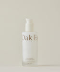 A frosted glass bottle with a white pump dispenser labeled Nourishing Cleansing Milk for sensitive skin stands against a plain, neutral background. The formula is enriched with Oat Amino Acids.