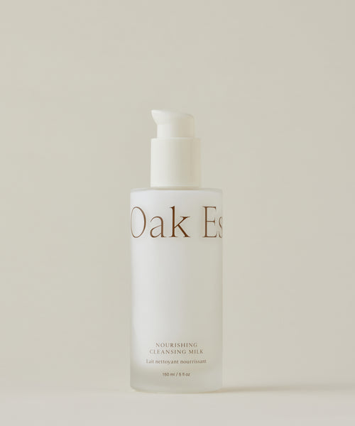 A frosted glass bottle with a white pump dispenser labeled Nourishing Cleansing Milk for sensitive skin stands against a plain, neutral background. The formula is enriched with Oat Amino Acids.