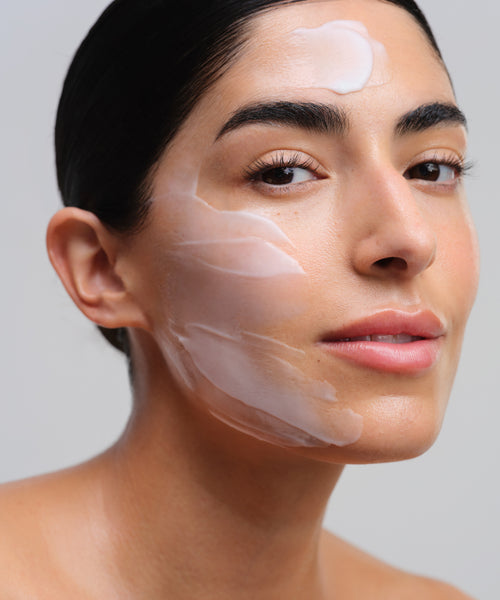 A woman with smooth, sensitive skin and dark hair pulled back has Nourishing Cleansing Milk applied to her forehead and cheeks, confidently facing the camera against a plain light background.