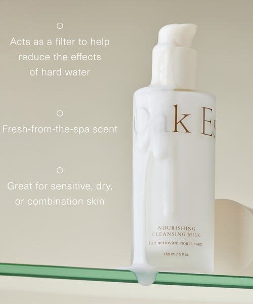 A white bottle of Nourishing Cleansing Milk rests on a glass shelf, some spilled product below. Text highlights its spa-like scent, suitability for sensitive or combination skin, and benefits for those with hard water.