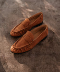 The Nubuck Dylan Loafer, a brown nubuck moccasin-style shoe with hand-stitched gathered edge details, is displayed on a textured taupe surface under soft natural light.
