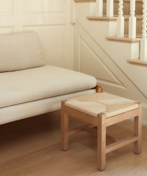 A minimalist living space with a beige sofa and wooden frame features the Montecito Stool, crafted from white oak and Danish paper cord, set against cream paneled walls and a staircase with spindle railings.