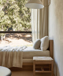 A cozy daybed with cream cushions and a knit blanket sits by a large window with sheer curtains and tree views; nearby, the Montecito Stool adds warmth, while soft light fills the serene room.