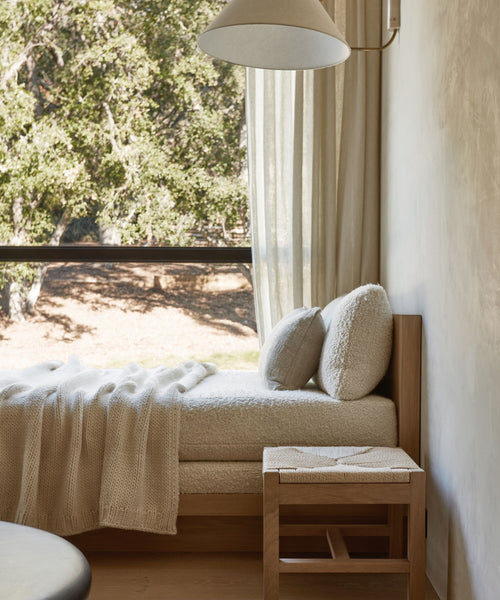 A cozy daybed with cream cushions and a knit blanket sits by a large window with sheer curtains and tree views; nearby, the Montecito Stool adds warmth, while soft light fills the serene room.