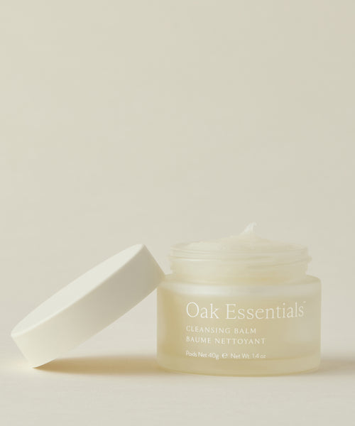 A frosted glass jar of Cleansing Balm with a white lid beside it displays the creamy formula inside, set against a light beige background.
