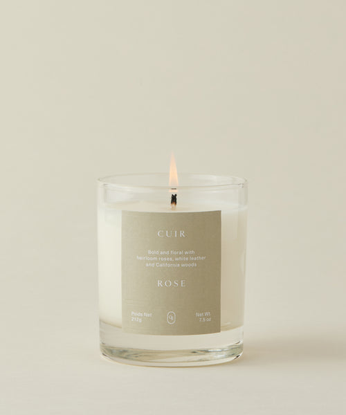 The Cuir Rose Candle glows in a clear glass jar with a beige label, filling your space with bold floral and woody notes of heirloom roses, white leather, and California woods.