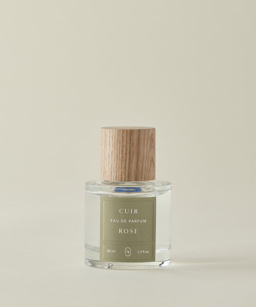 Against a beige backdrop, the clear glass Cuir Rose Eau De Parfum bottle—with a light wood-grain cap and green label—showcases its clean fragrance, blending floral and woody notes.