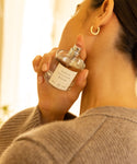 Wearing a beige sweater and gold hoop earrings, a woman sprays Santo Wood Eau De Parfum onto her neck.