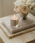 A Violet Santal Candle in a glass jar sits atop a closed book and Georgia O’Keeffe art book, next to white roses in a vase on a beige table, creating a calm and elegant scene with coastal nostalgia.