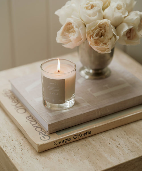 A Violet Santal Candle in a glass jar sits atop a closed book and Georgia O’Keeffe art book, next to white roses in a vase on a beige table, creating a calm and elegant scene with coastal nostalgia.