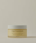 A frosted glass jar with a white lid labeled Moisture Rich Body Balm rests on a light beige background. The label, in English and French, highlights nourishing ingredients such as Cocoa Butter.