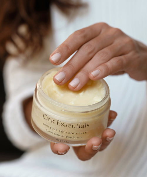A hand with manicured nails scoops creamy Moisture Rich Body Balm from a glass jar, while a softly blurred background draws focus to the product and the elegant gesture.