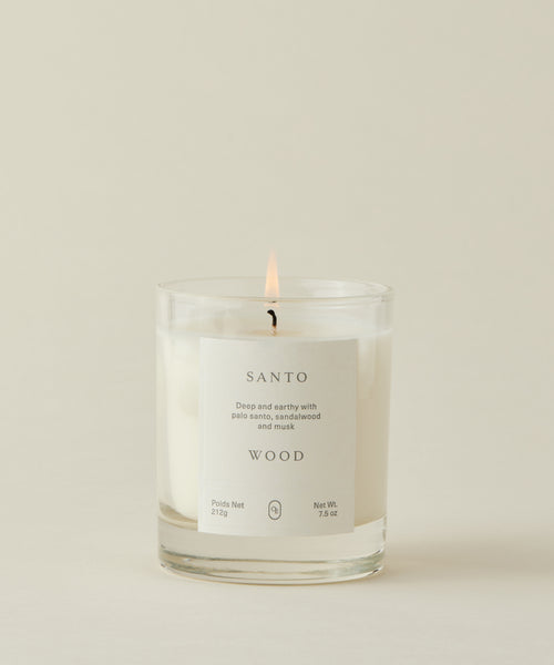 A lit Santo Wood Candle in a clear glass jar with a "SANTO WOOD" label sits against a light cream background. This musky candle blends deep palo santo, sandalwood, and musk. 212g / 7.5oz.