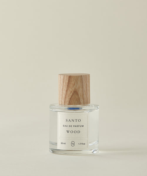 A clear glass bottle of Santo Wood Eau De Parfum with a wooden cap rests on a light beige surface. Its label shows 50 ml (1.7 fl oz). The clean, non-toxic fragrance stands out against the plain, light-colored background.