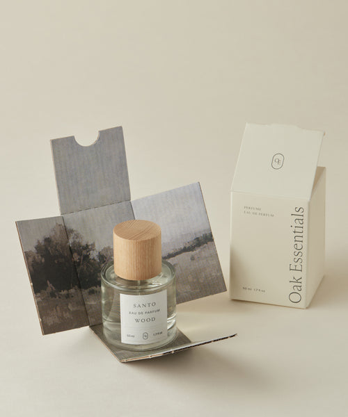 A glass bottle of Santo Wood Eau De Parfum with a wooden cap sits on its opened landscape-designed box, next to an upright cream-colored Oak Essentials perfume box.