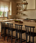 A warm, sunlit kitchen features wooden cabinets, open shelves with dishes, a large island with four black Hampton Counter Stools, a bowl of fruit, and a decorative light fixture hanging above.