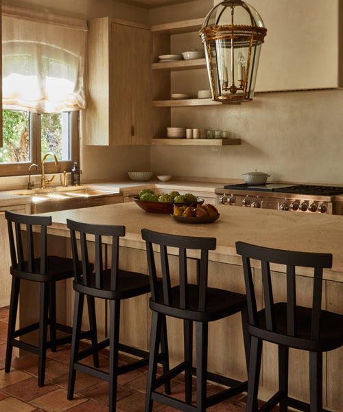 A warm, sunlit kitchen features wooden cabinets, open shelves with dishes, a large island with four black Hampton Counter Stools, a bowl of fruit, and a decorative light fixture hanging above.