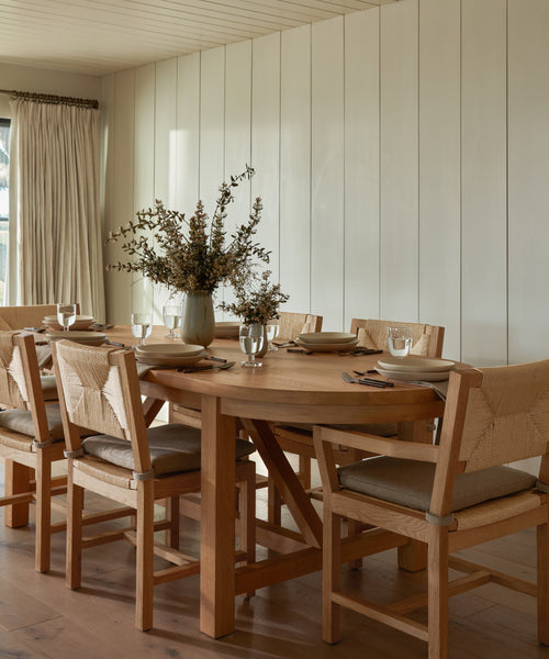 A wooden dining table set for six features the Montecito Dining Arm Chair, each with a Belgian linen cushion. Neutral dinnerware, glasses, and napkins enhance the vase centerpiece in a space with light wood walls and curtains.